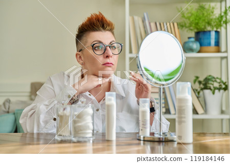 Middle aged woman looking in mirror, using face creams, cosmetic care products 119184146