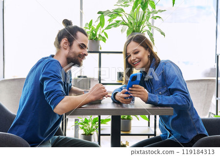 Young man woman sitting together in cafe coffee shop, talking, looking at smartphone Young man woman sitting together in cafe coffee shop, talking, looking at smartphone 119184313