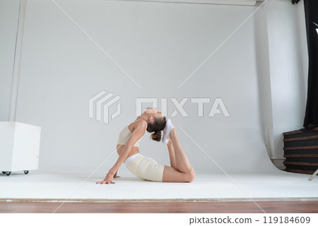 Dressed in a beige sports outfit, a woman bends backward in a graceful yoga pose against a plain white backdrop. This image captures the essence of yoga practice and the pursuit of a balanced 119184609
