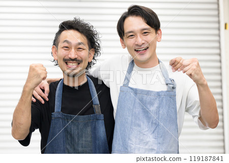 Two men wearing aprons, such as salespeople (clerks) at a recycling store or craftsmen, are friendly and look at the camera with their arms around each other's shoulders 119187841