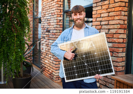 Portrait of bearded man holds solar panel, giving thumbs up, standing in front of brick building and lush greenery. Guy exudes calm and eco-conscious vibe, blending nature with urban elements. 119191384
