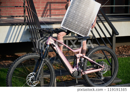 Cropped view of man cyclist holds solar panel for charging electric mountain bike. Guy in light gray shirt, blue shorts, and black helmet stands on staircase with brick wall in background. 119191385