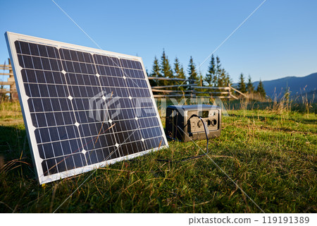 Tourist tent with solar panel and portable power station nearby in grassy field. Scene surrounded by rolling hills and wooden fence under clear blue sky, showcasing eco-friendly camping setup. 119191389