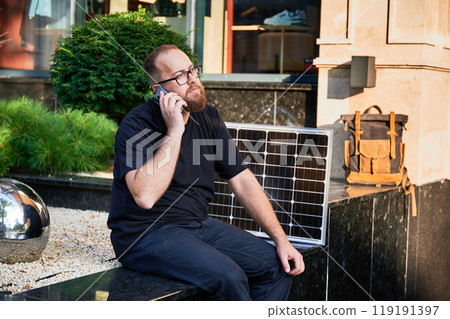 Bearded man talks on smartphone next to photovoltaic solar panel. Integration of sustainable renewable energy into everyday life, demonstrating practical use of solar power for charging devices. Bearded man talks on smartphone next to photovoltaic solar panel. Integration of sustainable renewable energy into everyday life, demonstrating practical use of solar power for charging devices. 119191397