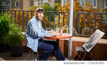 Bearded man works on laptop at outdoor round table, with glass of iced coffee. Solar panel nearby, emphasizing sustainable, modern eco-friendly workspace that combines technology and sustainability. Bearded man works on laptop at outdoor round table, with glass of iced coffee. Solar panel nearby, emphasizing sustainable, modern eco-friendly workspace that combines technology and sustainability. 119191401