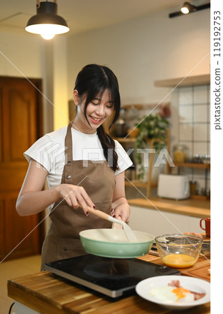 Cheerful young asian woman cooking scrambled eggs in frying pan on a stovetop 119192753