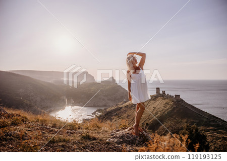 A blonde woman stands on a hill overlooking the ocean. She is wearing a white dress and she is enjoying the view. 119193125