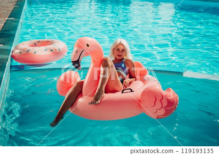 A woman is sitting on a pink inflatable flamingo in a pool. The flamingo is pink and has a blue stripe. The woman is smiling and she is enjoying herself. Concept of fun and relaxation. A woman is sitting on a pink inflatable flamingo in a pool. The flamingo is pink and has a blue stripe. The woman is smiling and she is enjoying herself. Concept of fun and relaxation. 119193135