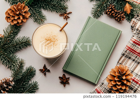 Green recipe book next to creamy eggnog drink with spices. Pinecones and holiday greenery create festive atmosphere. Green recipe book next to creamy eggnog drink with spices. Pinecones and holiday greenery create festive atmosphere. 119194476