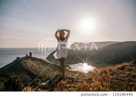 A blonde woman stands on a hill overlooking the ocean. She is wearing a white dress and she is enjoying the view. 119196440