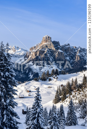 Sassongher mountain rises above a snow covered valley in the Dolomites with wooden cabins 119196599