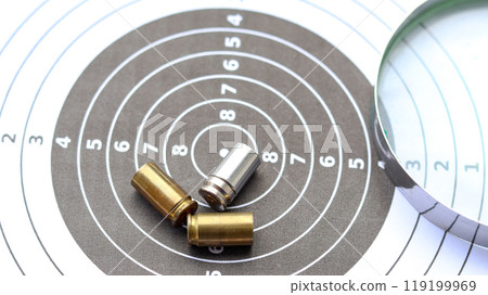 Empty bullet casing on a shooting target, with a part of magnifying glass, evoking the concept of crime scene investigation and unraveling mysteries. Empty bullet casing on a shooting target, with a part of magnifying glass, evoking the concept of crime scene investigation and unraveling mysteries. 119199969