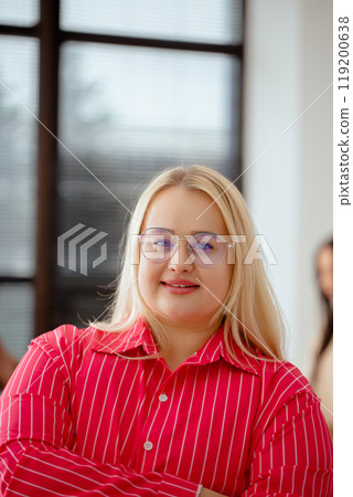 Woman in a pink shirt stands in front of a window with her arms crossed, people in the team work in the background Woman in a pink shirt stands in front of a window with her arms crossed, people in the team work in the background 119200638