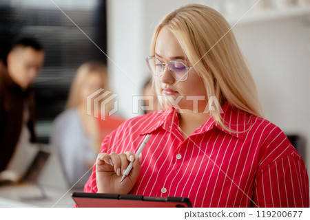 Woman in a pink shirt is writing on a tablet. She is wearing glasses and has long blonde hair Woman in a pink shirt is writing on a tablet. She is wearing glasses and has long blonde hair 119200677