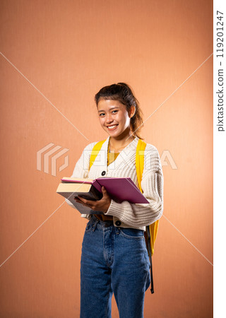 A Cheerful Student Holding Several Books with a Backpack Against a Warm Background 119201247
