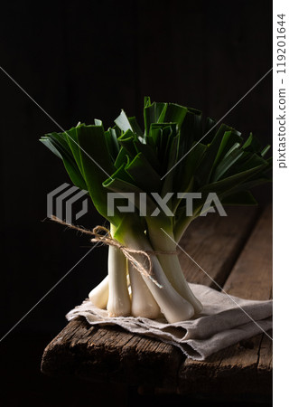 A bunch of leeks stands on the corner of the table on a dark background 119201644