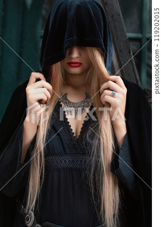 Gloomy blonde woman in black dress, vintage Scythian necklace and hood,against the background of an old hut Gloomy blonde woman in black dress, vintage Scythian necklace and hood,against the background of an old hut 119202015