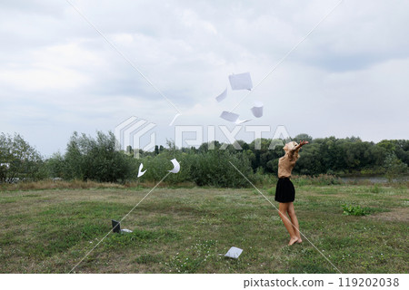 Woman is standing in a field with a laptop open and papers flying around her 119202038