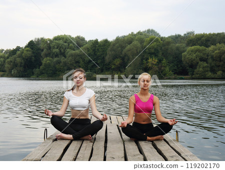 Two teenage girls doing yoga exercises for concentration and relaxation on a wooden pier 119202170