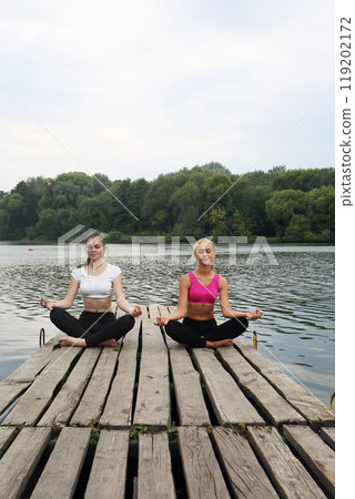 Two teenage girls doing yoga exercises for concentration and relaxation on a pier by the river Two teenage girls doing yoga exercises for concentration and relaxation on a pier by the river 119202172