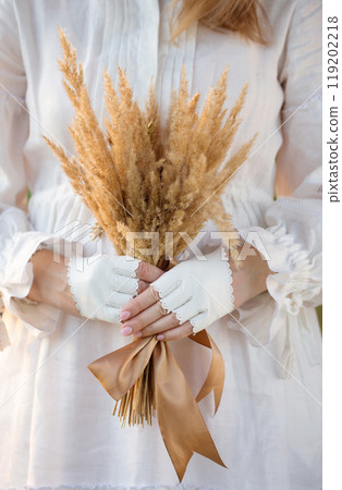 A woman in a white vintage dress and white leather mittens holds a bouquet of dried flowers, close-up A woman in a white vintage dress and white leather mittens holds a bouquet of dried flowers, close-up 119202218