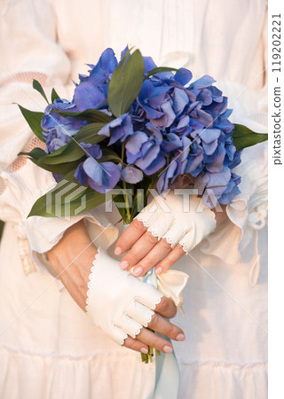 A woman in a white vintage dress and white leather mittens holds a bouquet of blue hydrangea, close-up 119202221