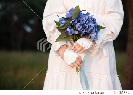 A woman in a white vintage dress and white mittens holds a bouquet of blue hydrangea, close-up A woman in a white vintage dress and white mittens holds a bouquet of blue hydrangea, close-up 119202222