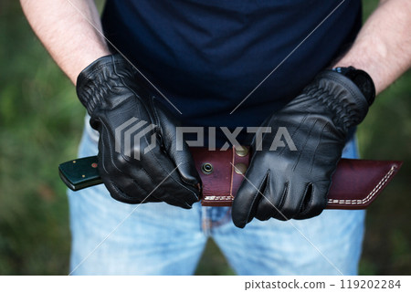 A man in black gloves holds a large knife and a leather cover for the knife A man in black gloves holds a large knife and a leather cover for the knife 119202284