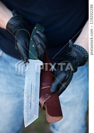 A man in black leather gloves holds a large knife and a leather cover for the knife 119202286