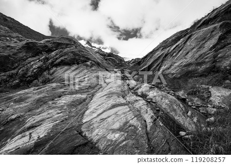A rugged hiking path ascends through the rocky landscape of Schlaten Glacier in Austria, surrounded by towering peaks and dramatic cloud cover. Black and white photography. 119208257
