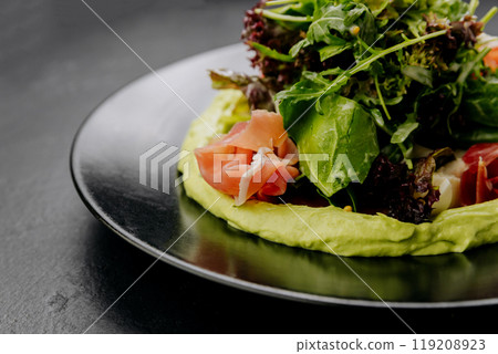 Fresh salad with avocado cream and prosciutto, served on a black plate, perfect for gourmet dining Fresh salad with avocado cream and prosciutto, served on a black plate, perfect for gourmet dining 119208923