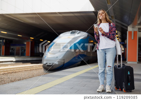 A Young Woman Standing at a Busy Train Station with Her Luggage and Smartphone in Hand A Young Woman Standing at a Busy Train Station with Her Luggage and Smartphone in Hand 119209385