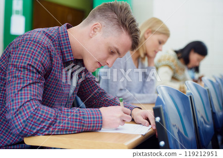 University student, desk and writing exam in classroom for education assessment for knowledge, diploma or scholarship. Men, woman and test in London lecture hall or study document, academy or college 119212951