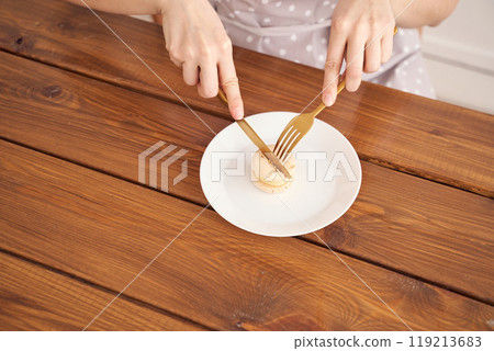 Minimalist dining: hands cutting macaron with fork and knife on wooden table Minimalist dining: hands cutting macaron with fork and knife on wooden table 119213683