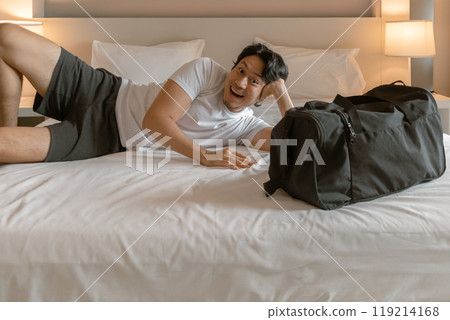 Happy traveler man lying on the hotel bed with luggage travel bag. 119214168