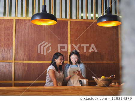 Restaurant, tablet and asian women at counter together checking sales, booking or menu for small business. Social media, content marketing and startup cafe owner with waitress scroll on digital app. 119214379