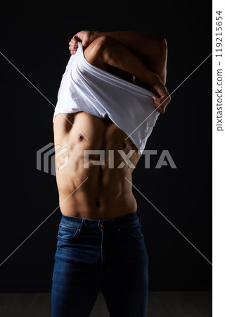 Fitness, undressing and muscle with man in studio for strong, body and health. Sports, wellness and workout with person and remove tshirt on dark background for fashion, gym and athlete motivation 119215654