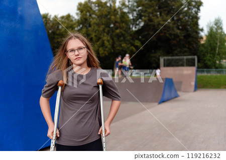 Young girl standing leaning on crutches on the background of skate park, gray T-shirt with mockup for advertising 119216232