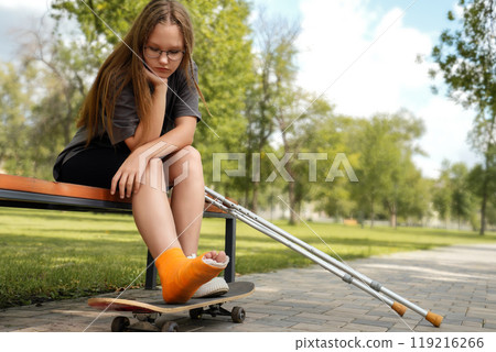 sad young girl sits with a broken leg in a park on a bench and looks sadly at a skateboard board. Summer injuries, medicine 119216266