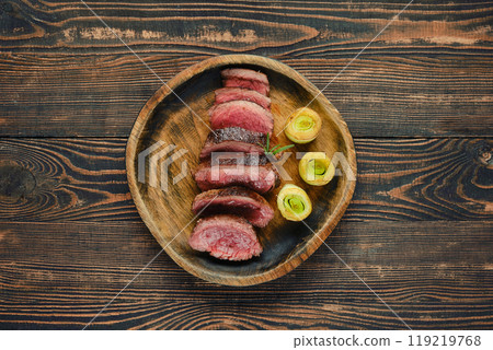 Sliced roast beef steak with roasted leek served on a wooden plate against a rustic background 119219768