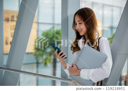 Professional woman in white shirt multitasking with laptop and phone in airport. Productive work on-the-go. Stay connected and productive while traveling 119219951