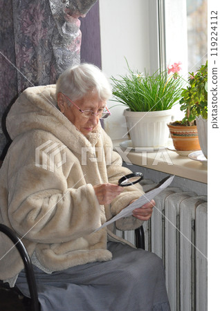 The senior woman holding gas bill in front of heating radiator. Payment for heating in winter. Freezing old people in Europe. The senior woman at home. 119224112