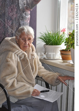 The senior woman holding gas bill in front of heating radiator. Payment for heating in winter. Freezing old people in Europe. The senior woman at home. 119224114