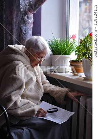 The senior woman holding gas bill in front of heating radiator. Payment for heating in winter. Freezing old people in Europe. The senior woman at home. The senior woman holding gas bill in front of heating radiator. Payment for heating in winter. Freezing old people in Europe. The senior woman at home. 119224115