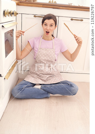 Cheerful woman in apron sitting on kitchen floor holding cooking utensils Cheerful woman in apron sitting on kitchen floor holding cooking utensils 119226797