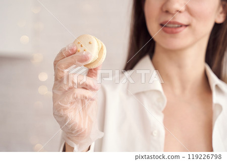 Smiling woman in white shirt holding delicious macaron with gloved hand 119226798