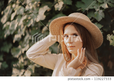 Woman with straw hat stands in front of vineyard. She is wearing a light dress and posing for a photo. Travel concept to different countries Woman with straw hat stands in front of vineyard. She is wearing a light dress and posing for a photo. Travel concept to different countries 119227333