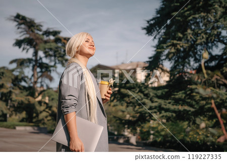 A woman is walking down the street with a white laptop bag and a cup of coffee. She is smiling and enjoying the sunny day. A woman is walking down the street with a white laptop bag and a cup of coffee. She is smiling and enjoying the sunny day. 119227335