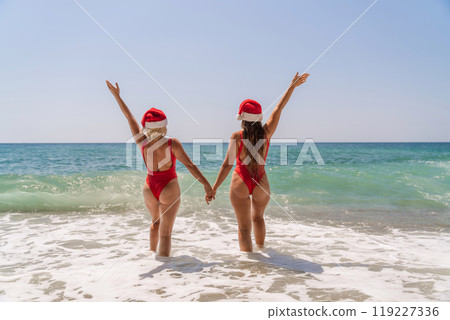 Women Santa hats ocean play. Seaside, beach daytime, enjoying beach fun. Two women in red swimsuits and Santa hats are enjoying themselves in the ocean waves and raising their hands up. Women Santa hats ocean play. Seaside, beach daytime, enjoying beach fun. Two women in red swimsuits and Santa hats are enjoying themselves in the ocean waves and raising their hands up. 119227336