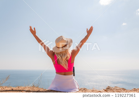A woman wearing a pink top and a straw hat is sitting on a beach, looking out at the ocean. She is smiling and she is enjoying the view. A woman wearing a pink top and a straw hat is sitting on a beach, looking out at the ocean. She is smiling and she is enjoying the view. 119227361
